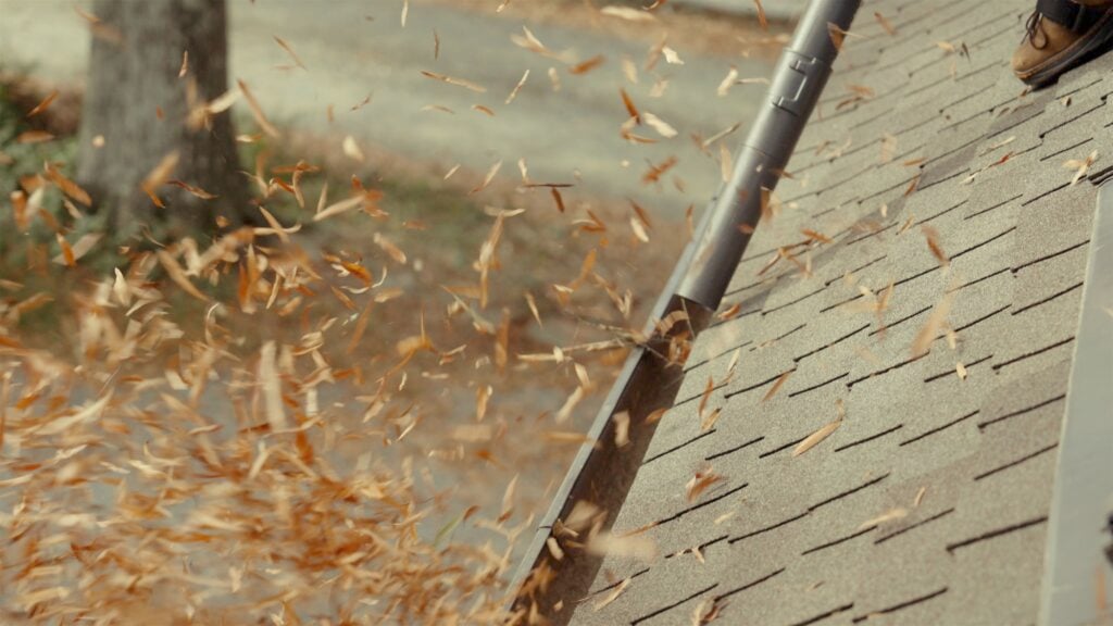 A person uses a leaf blower for roof cleaning, expertly clearing fallen leaves from the asphalt shingles. The action sends a flurry of dry leaves swirling into the air, with a tree and path visible in the background.