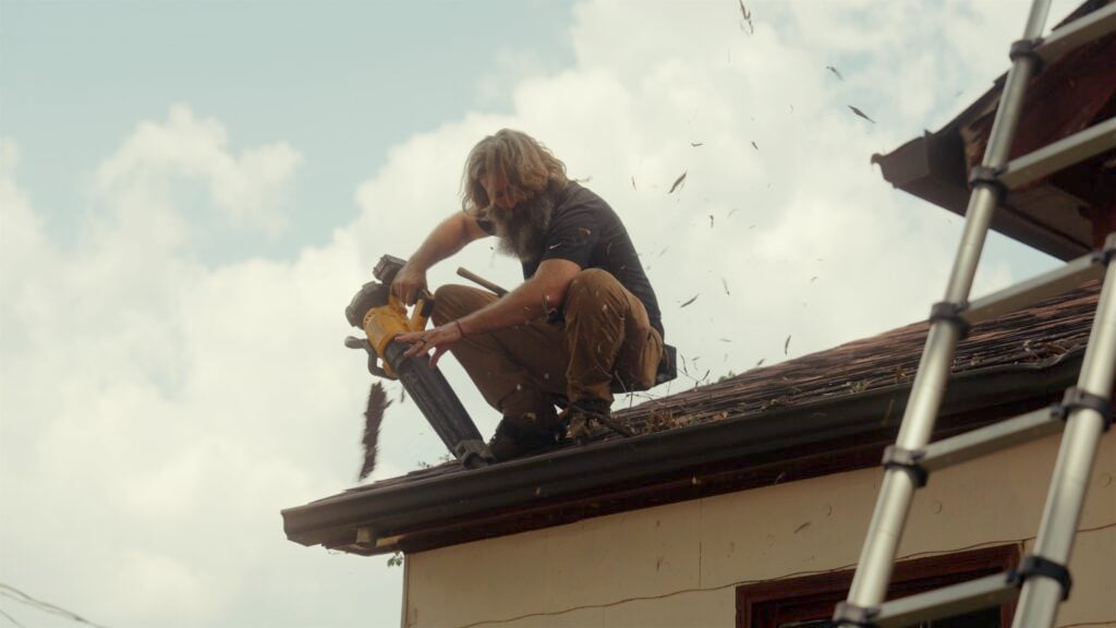 A person with long hair and a beard conducts roof cleaning using a leaf blower to clear debris from a rooftop. They're crouched near the edge, dressed casually. A ladder is visible in the foreground against a partly cloudy sky.