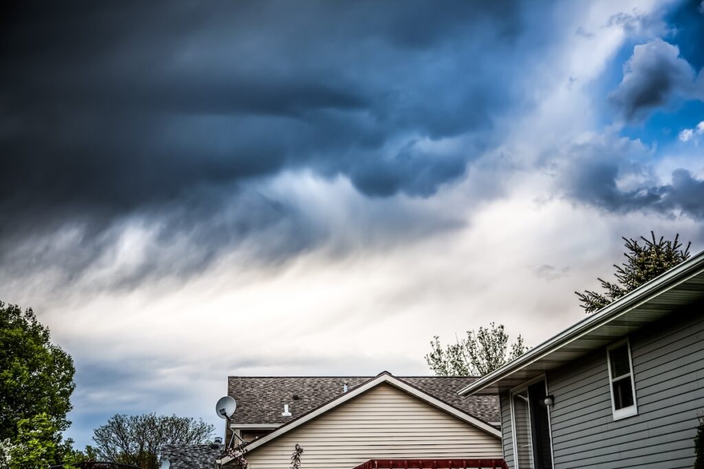 Dark storm clouds hover over suburban rooftops, contrasting with lighter clouds. The scene includes trees and various rooflines of houses in need of repair, creating a dramatic atmosphere that hints at potential storm damage.
