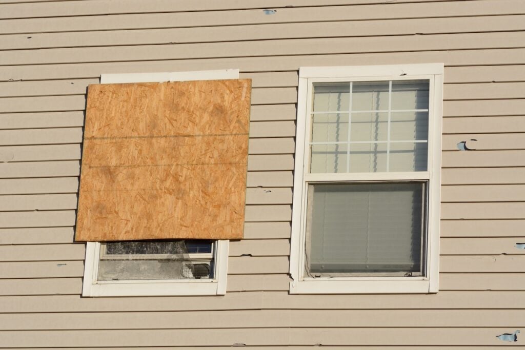 A beige house exterior features two windows; the left one is shielded by a large piece of plywood, indicating recent storm damage