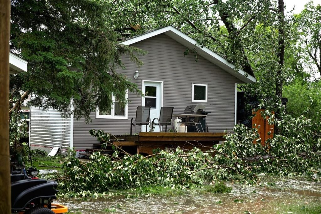 A small gray house with white trim is surrounded by fallen tree branches and debris, revealing signs of storm damage. A wooden porch holds patio chairs and a barbecue grill. The yard is flooded, while trees in the background emphasize the need for roof repair at this apex of weather's wrath.
