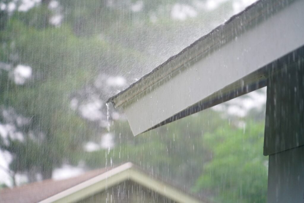 Rain pours from a sloped roof, hinting at the need for roof repair as droplets cascade over the edge. In the background, blurry greenery and other buildings form an apex-like silhouette, adding depth and context to this scene of potential storm damage.