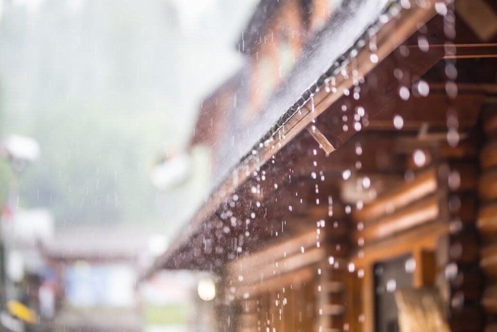 Raindrops gently cascade from the edge of a sloped wooden roof in Chapel Hill, with blurred greenery in the background. The log-built structure stands tranquil amid rainfall, contrasting sharply with memories of past storm damage and the repairs that followed.
