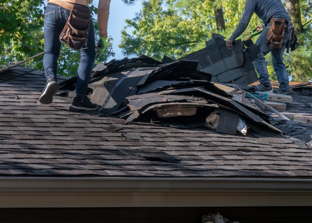 Two workers with tool belts stand on a house roof in Durham, expertly handling storm damage repair. They are removing old shingles, which are piled up on the roof. Green trees are visible in the background against a clear sky.