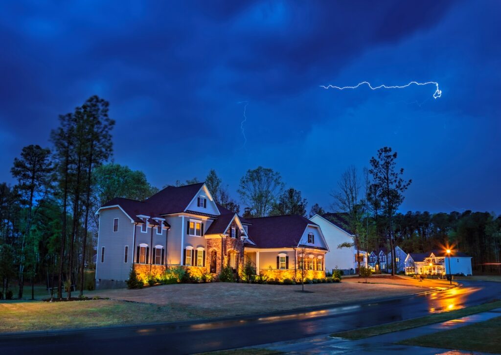 A large house with warm exterior lighting sits under a dark, cloudy sky in Cary. Lightning illuminates the night sky, hinting at potential storm damage. The wet pavement reflects the lights, and trees frame the scene, adding a touch of eerie beauty to the impending need for roof repair.