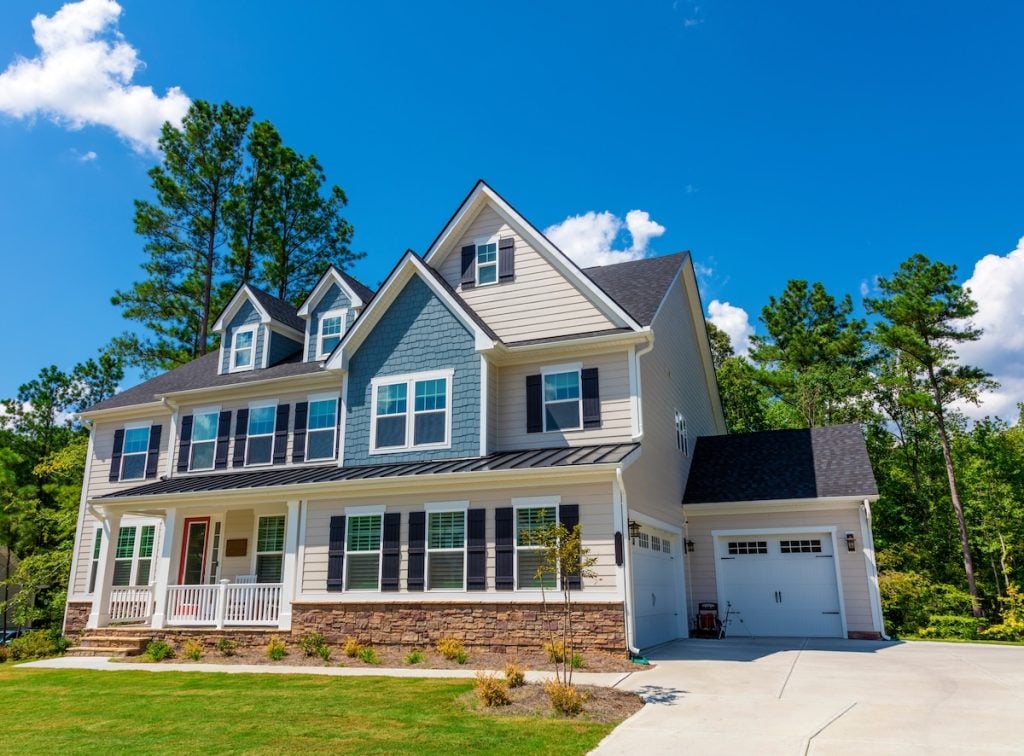 A large, modern two-story house with light siding and newly completed roof upgrades features dark roof shingles, dormer windows, and a front porch.