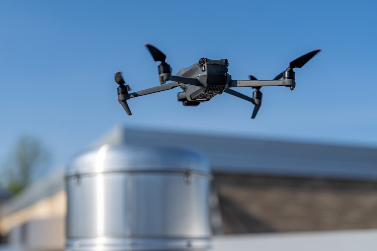 A black drone with four propellers flies above a building rooftop with a shiny silver vent, conducting a roof inspection