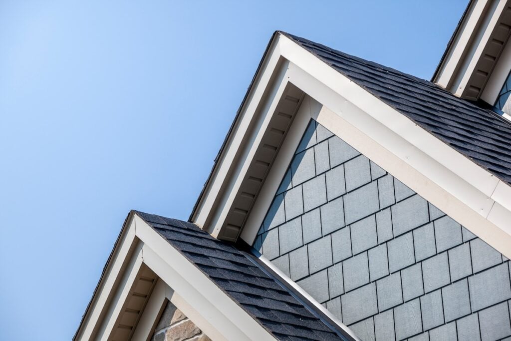 Close-up of a modern house gable roof with slate shingles and white trim against a clear blue sky. The roof has multiple peaks, and the siding features different shades of gray for a textured appearance.