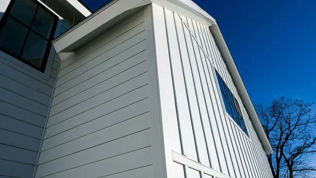Close-up of a modern white board and batten house exterior with a large window. The deep blue sky contrasts with the building's clean lines. Leafless tree branches are visible in the background.