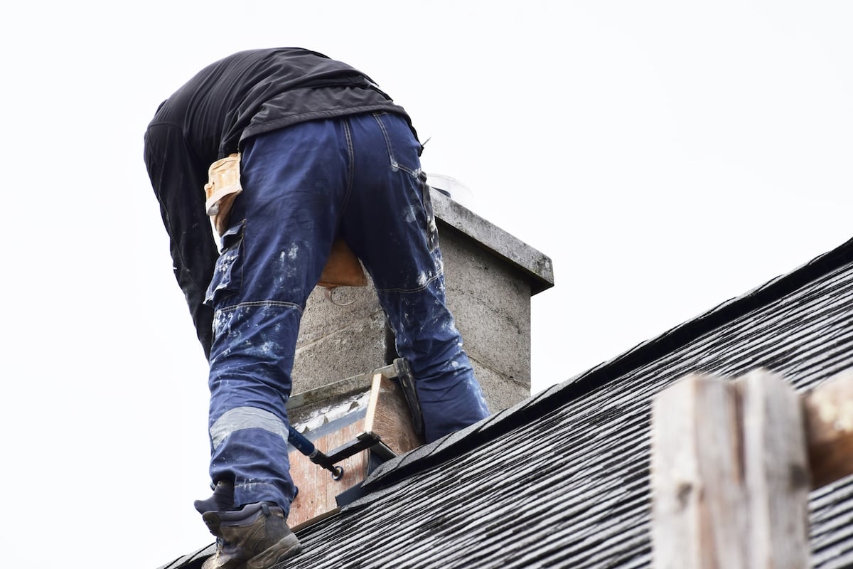A person in work clothes and a tool belt is standing on a sloped roof, performing chimney leak repair
