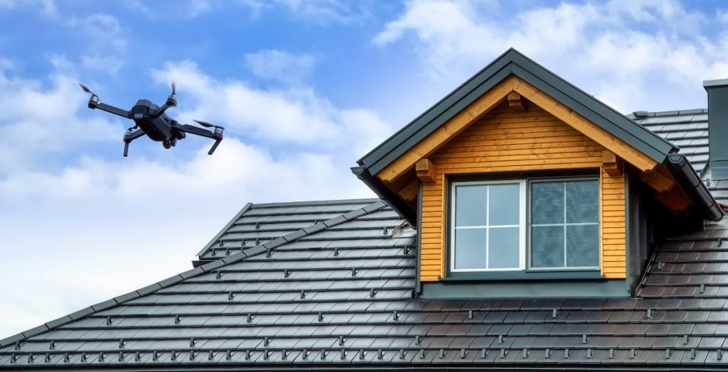 A black drone conducts a roof inspection near the house’s gray-tiled roof and wooden dormer window