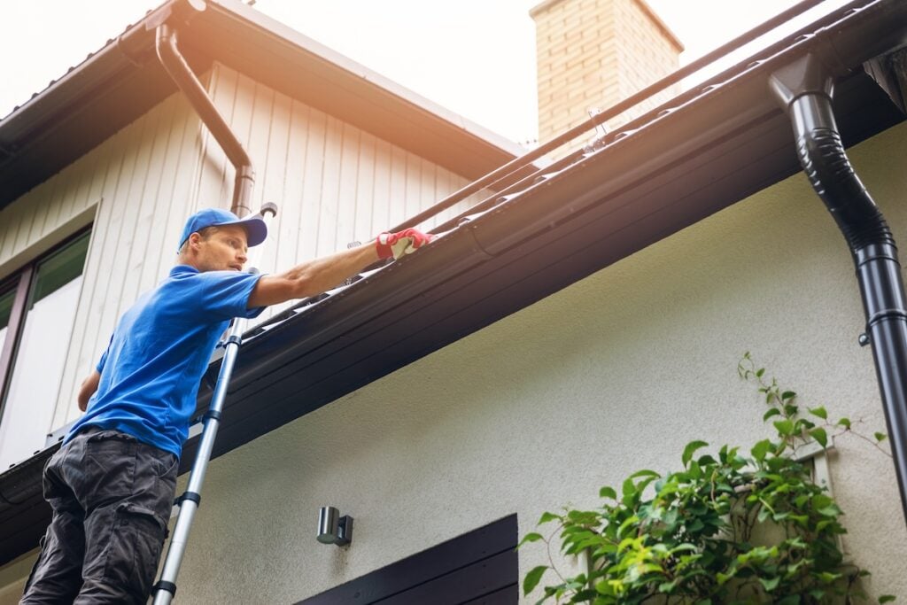 A person wearing a blue shirt and cap is standing on a ladder, cleaning the gutter of a house. They are using gloves for safety. The house has light-colored walls and a chimney is visible in the background. Green plants are in the foreground.