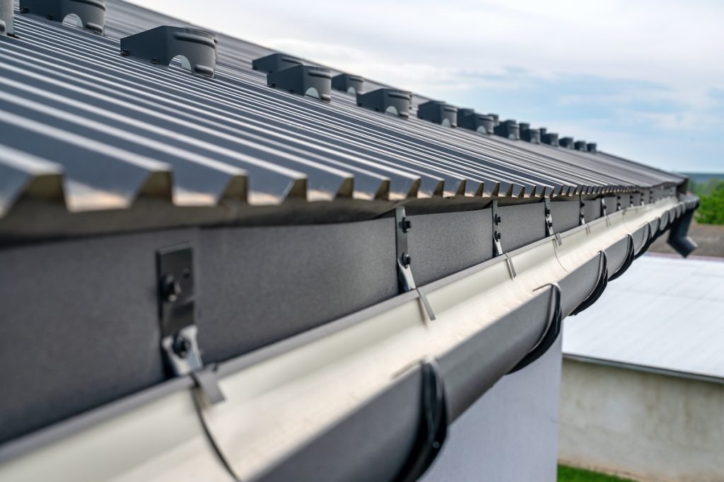 Close-up view of a gray metal roof with ridges and an attached rain gutter system, showcasing recent roof upgrades on a modern building.