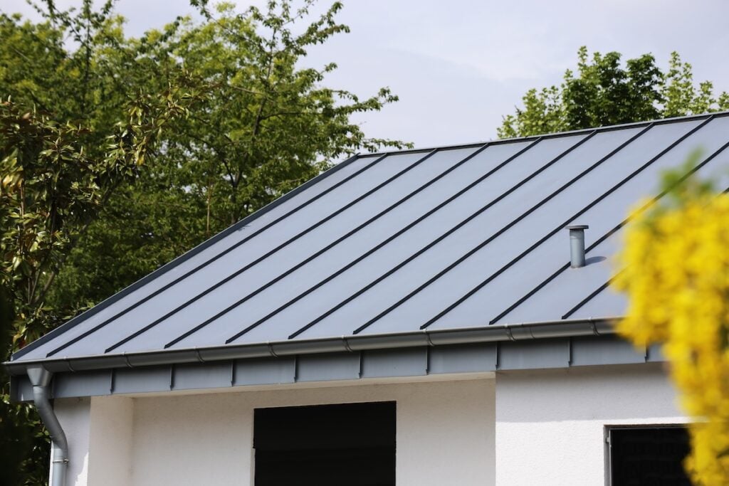 A modern house with a sleek metal roof is surrounded by trees. The roof features clean lines and a smokestack.