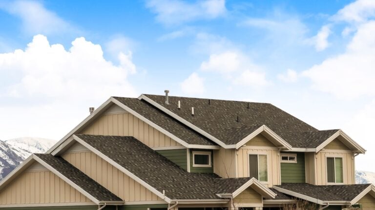 A suburban house with green and beige siding and a multi-gabled roof against a blue sky with clouds.