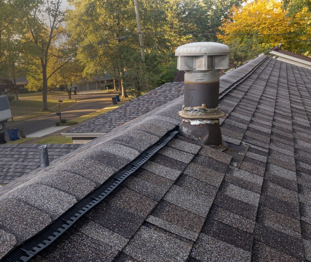 A rooftop with brown shingles features a metal vent pipe and a black ridge vent