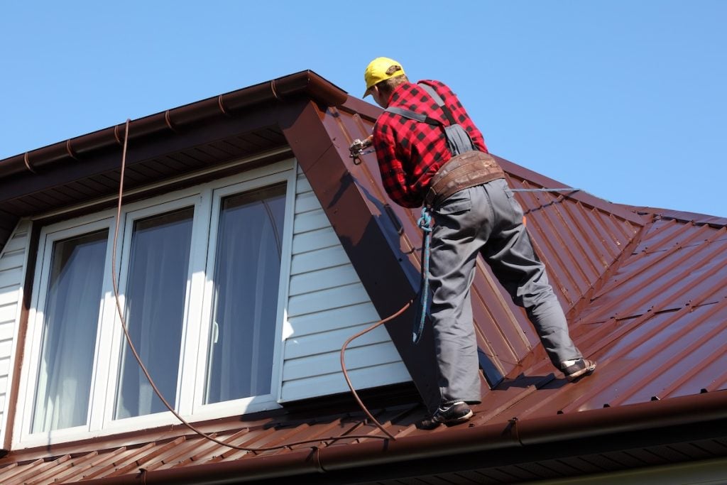 A worker in overalls and a yellow cap stands on a brown roof, applying metal roof coating with a spray gun