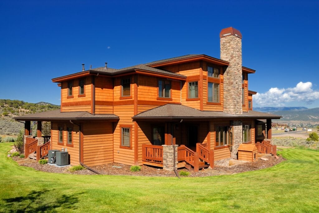 A large, modern, two-story wooden house with a stone chimney and wraparound porch
