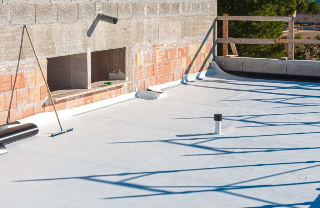 A flat roof under construction with a white waterproof membrane being installed, surrounded by unfinished brick and concrete walls, tools, and wooden safety railings—highlighting factors that influence flat roof replacement cost.