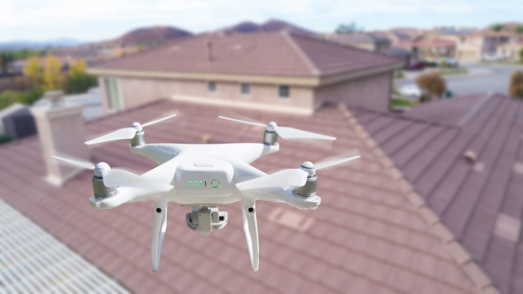 A white quadcopter drone with a camera hovers in the air above the roof of a suburban house, performing a drone roof inspection