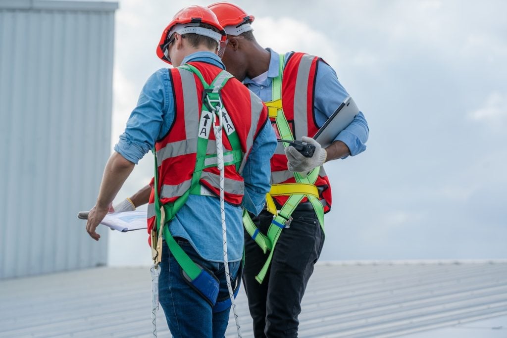 Two construction workers wearing safety harnesses, helmets, and reflective vests stand on a metal rooftop during a commercial roof inspection, looking at documents and a tablet