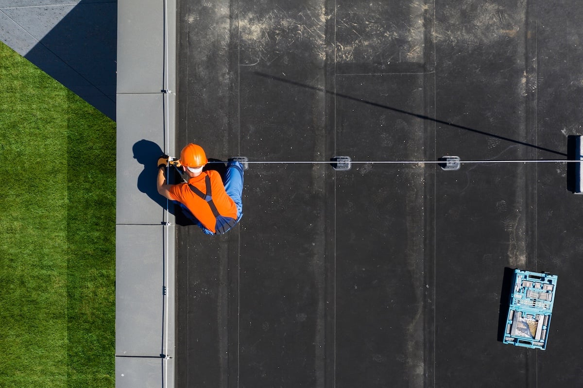 A worker is kneeling on a flat rooftop, appearing to perform a commercial roof inspection or install equipment.