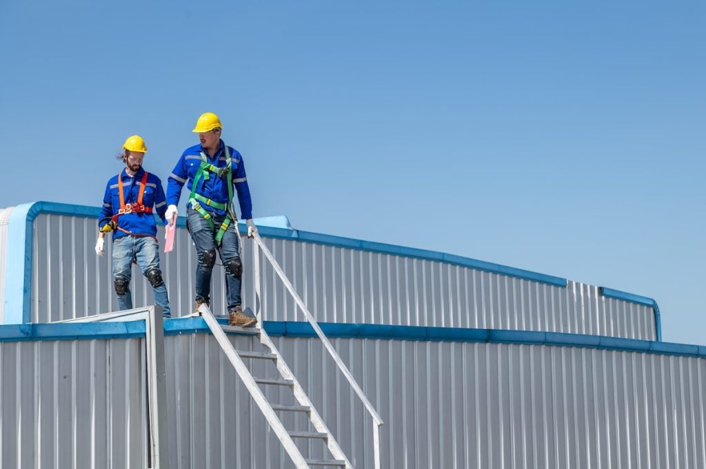 Two construction workers wearing safety gear and helmets stand on stairs atop a large, curved metal structure, performing a commercial roof inspection