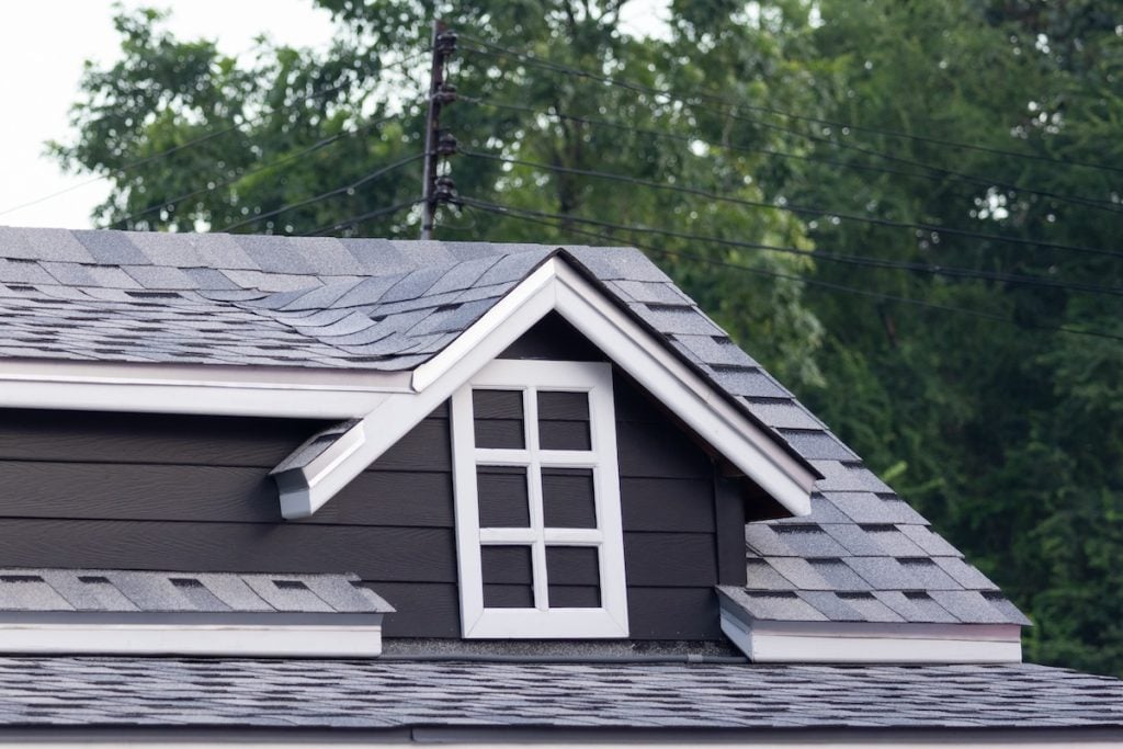 A close-up of a house roof with gray dimensional shingles, a small dormer window with white trim