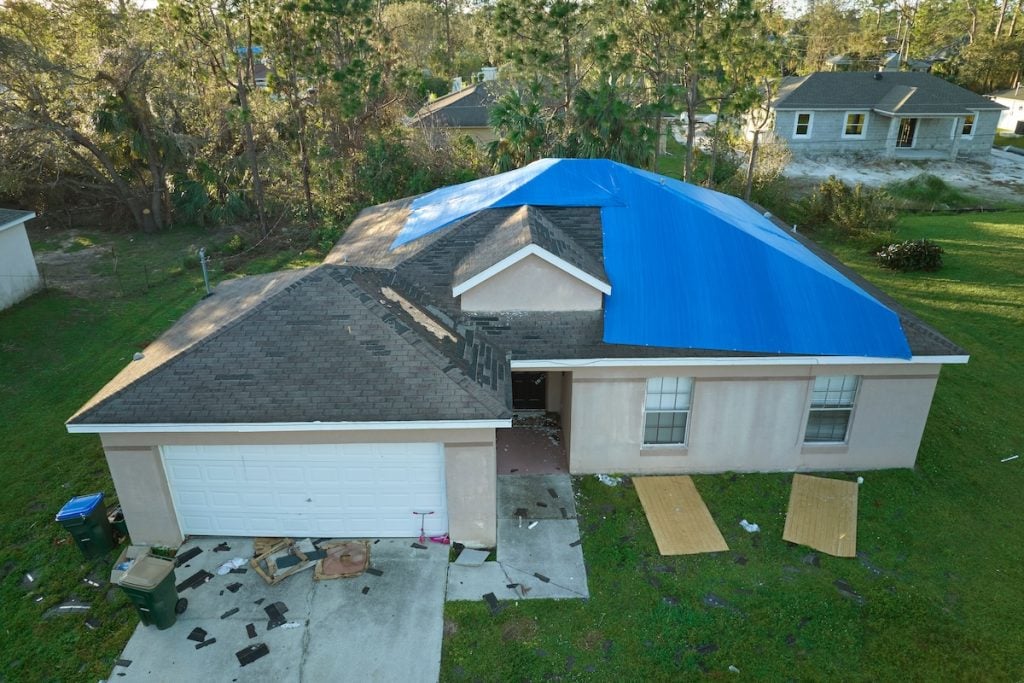A single-story house with an emergency roof tarp covering part of its roof, likely due to storm damage. Debris and wooden boards are scattered on the driveway and lawn