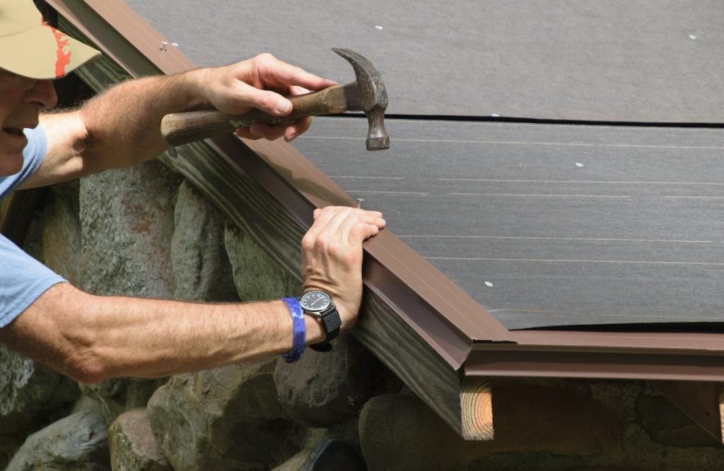 A person uses a hammer to drive a nail into the roof edge, securing dark roofing material and flashing on roof edge above a stone wall.