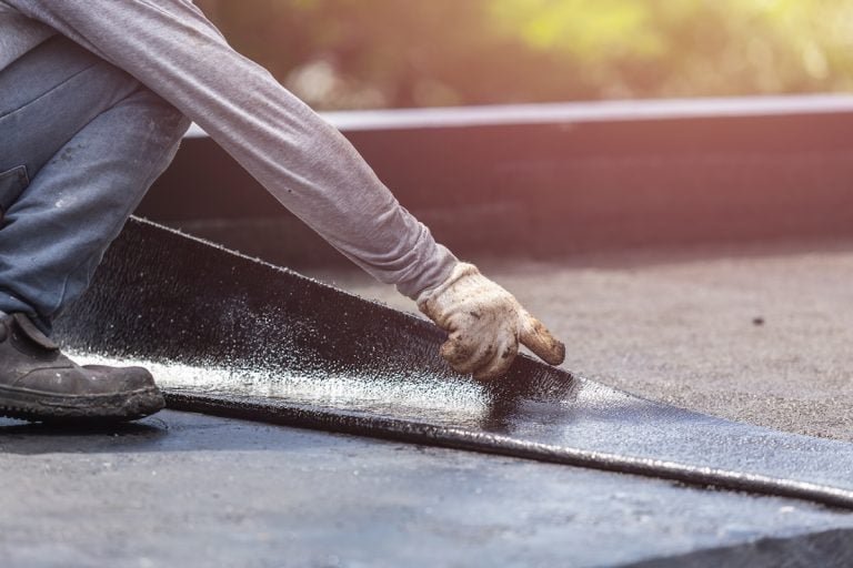 A person wearing gloves and a long-sleeve shirt is installing a sheet of roofing material on a flat roof
