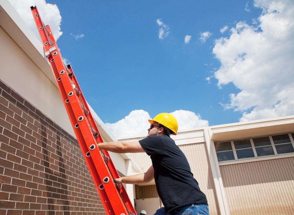 A person wearing a yellow hard hat and black shirt climbs an orange extension ladder positioned against a brick building, preparing for a commercial roof inspection