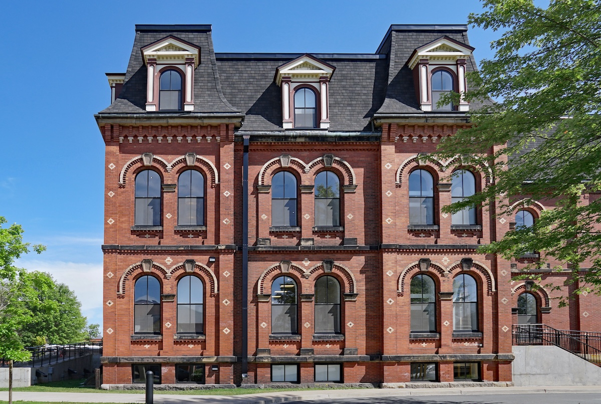 A historic red brick building with tall arched windows, decorative stone accents, and a slate mansard roof under a clear blue sky