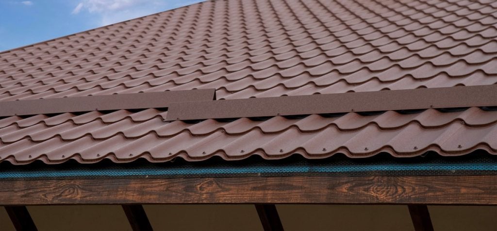 Close-up view of a brown metal tile roof with a wavy pattern. A piece of metal flashing sits on roof waiting to be installed