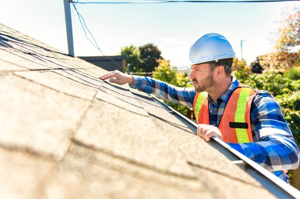A man in a hard hat and safety vest inspects the shingles on a house roof