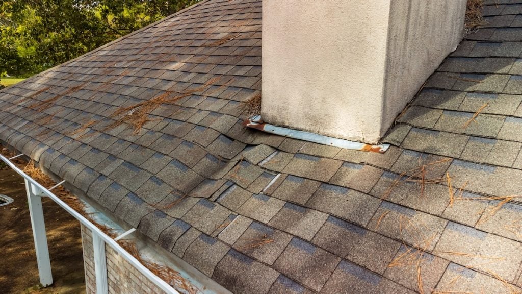 A roof with gray shingles shows visible damage and sagging near a chimney. Some shingles and flashing on the roof edge are missing