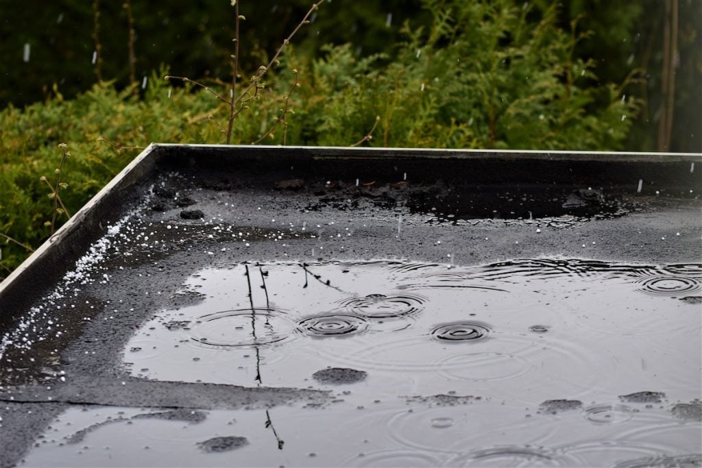 Rain falls on a flat black rooftop, creating ripples and small splashes in puddled water