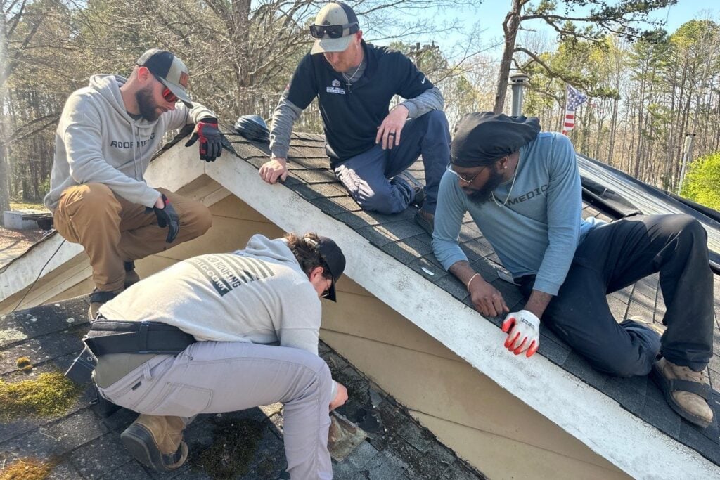 black shingles Four men in work clothes and gloves kneel on a house roof covered with black shingles, inspecting or repairing an area near the edge; trees and an American flag are visible in the background. The photo is dated March 20, 2024.