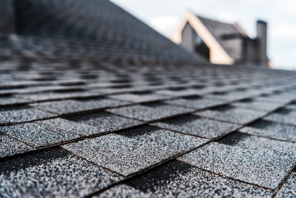 Close-up of gray and black shingles on an asphalt roof, with a blurred background showing part of a house roof and chimney against a cloudy sky.