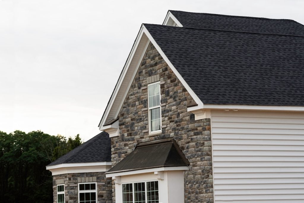 A close-up view of a modern house exterior featuring gray stonework, white siding, white trim, and black shingles roofing