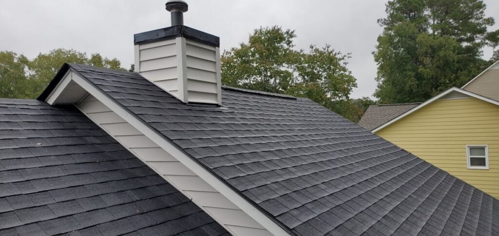 black shingles A close-up view of a house roof with black shingles, a white chimney, and trees and houses in the background on a cloudy day.