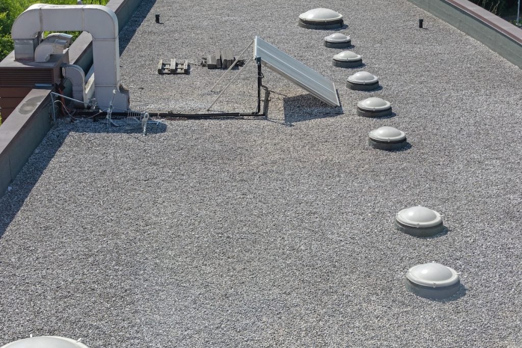 A flat tar and gravel roof with several round skylights, HVAC equipment, and a single tilted solar panel. The roof is bordered by low parapet walls, with some visible cables and pipes running along its surface.
