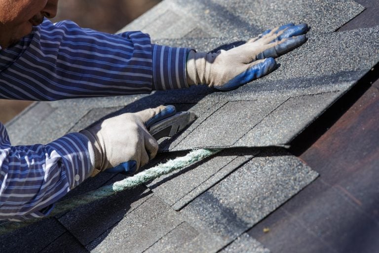 A person wearing gloves and a striped shirt cuts asphalt roofing shingles with a utility knife while working on a roof.