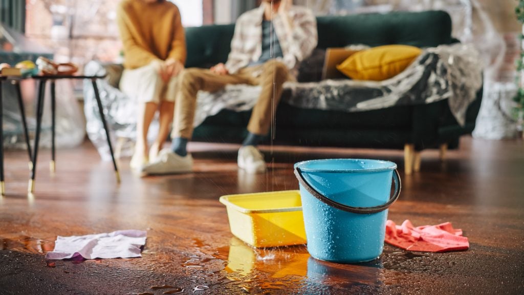 A blue bucket and a yellow basin catch water dripping from a ceiling leak, possibly caused by damaged step flashing roofing, onto a wooden floor; towels are nearby as two concerned people sit on a couch in the blurry background.