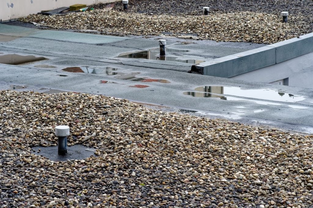 A flat tar and gravel roof covered with small stones, several vents, and puddles of water