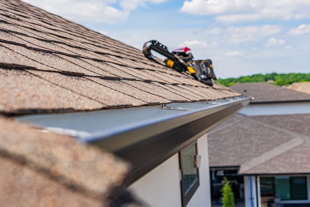 Close-up of a house roof with brown shingles and a black rain gutter, highlighting how often gutters should be cleaned. A yellow and black tool bag sits on the rooftop. Other houses and greenery are visible under a partly cloudy sky.