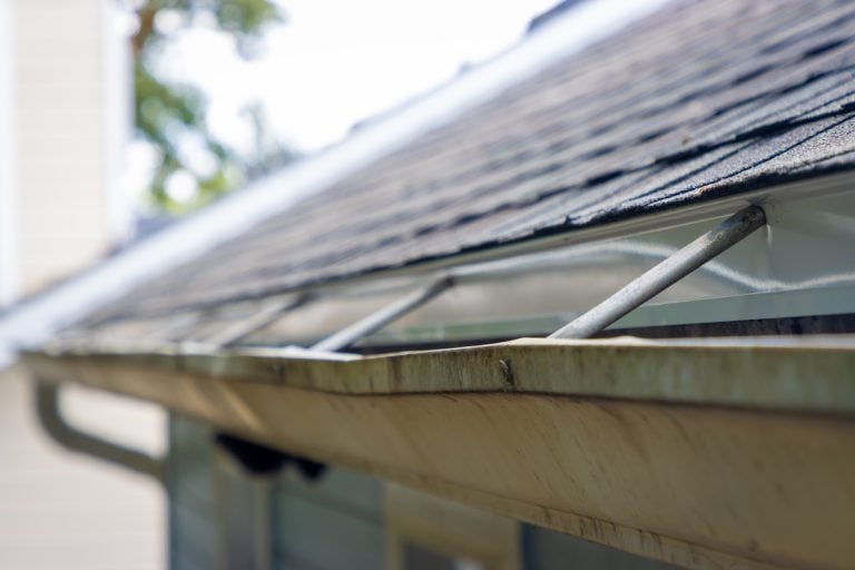 Close-up of a house’s roof with a dirty, clogged rain gutter and metal braces, showing grime buildup along the edge and shingles above—highlighting the need to address issues like how to fix sagging gutters.