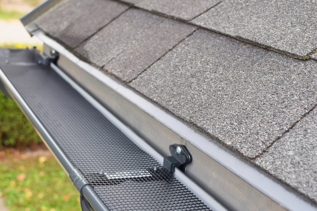 A close-up view of a house roof with asphalt shingles and a metal mesh gutter guard installed along the roof edge, ideal for those looking to prevent clogs and learn how to fix sagging gutters.