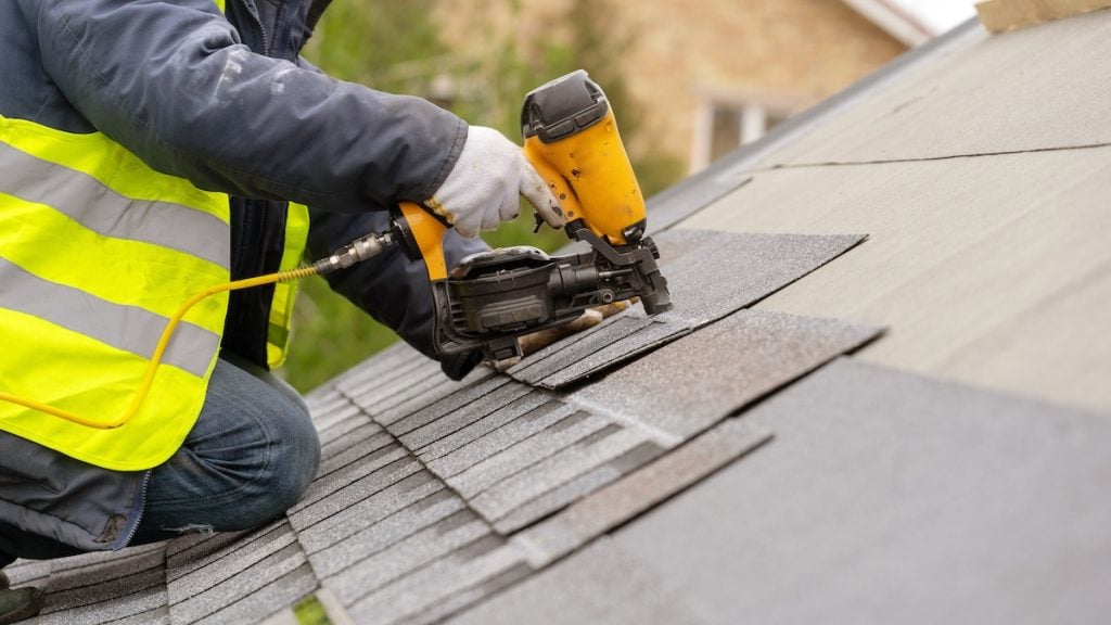 A worker in a yellow safety vest uses a nail gun to install asphalt shingles on a roof.