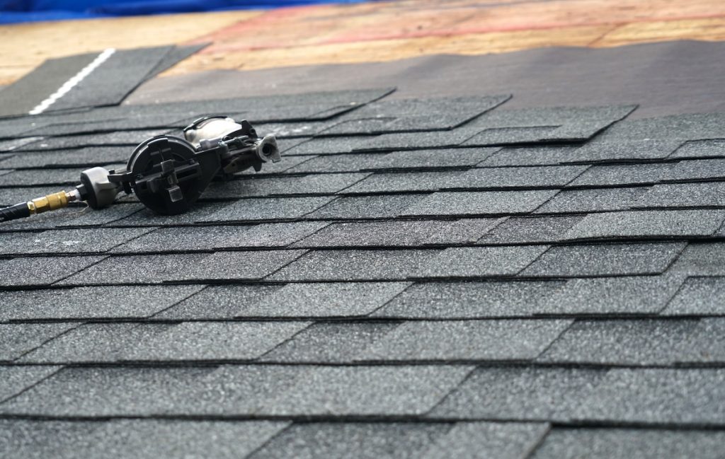 A close-up of gray asphalt shingles being installed on a roof, with a pneumatic nail gun resting on the shingles. The background shows partially completed roofing layers—an important scene to consider when learning how to choose a roofing contractor.